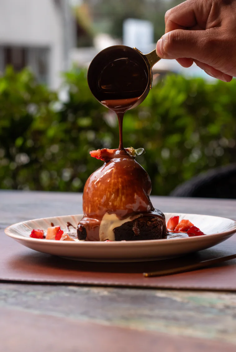 Calda de chocolate quente sendo derramada sobre uma bola de sorvete em cima de um brownie, com morangos picados no prato.