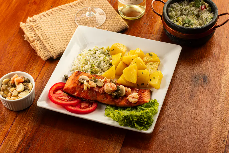 Filé de salmão grelhado em prato quadrado branco, com batatas douradas, arroz verde, camarões, alcaparras e salada.