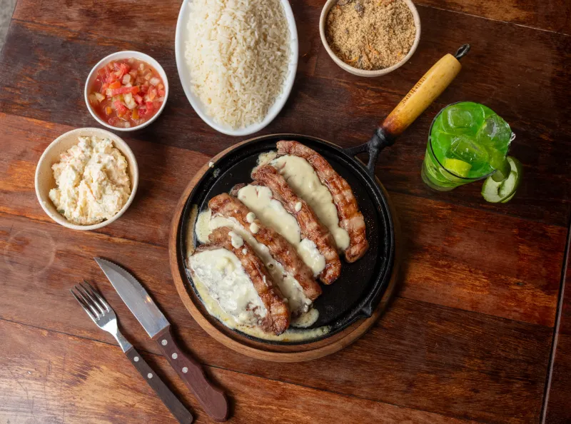 Picanha fatiada com creme de queijo em chapa de ferro, vista de cima, com arroz, farofa e vinagrete.