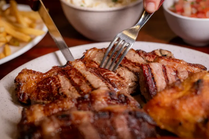 Garfo e faca cortando um churrasco misto grelhado, com fritas e vinagrete ao fundo.