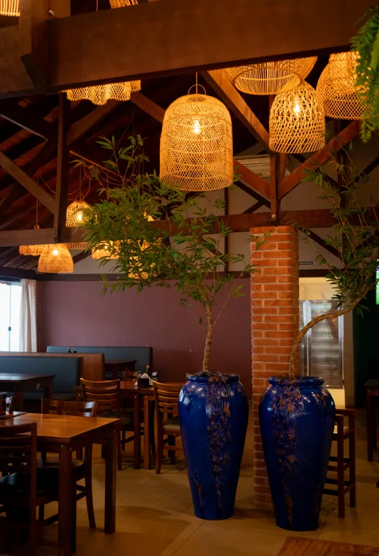 Salão de restaurante com mesas de madeira, luminárias de palha e pilar de tijolos.